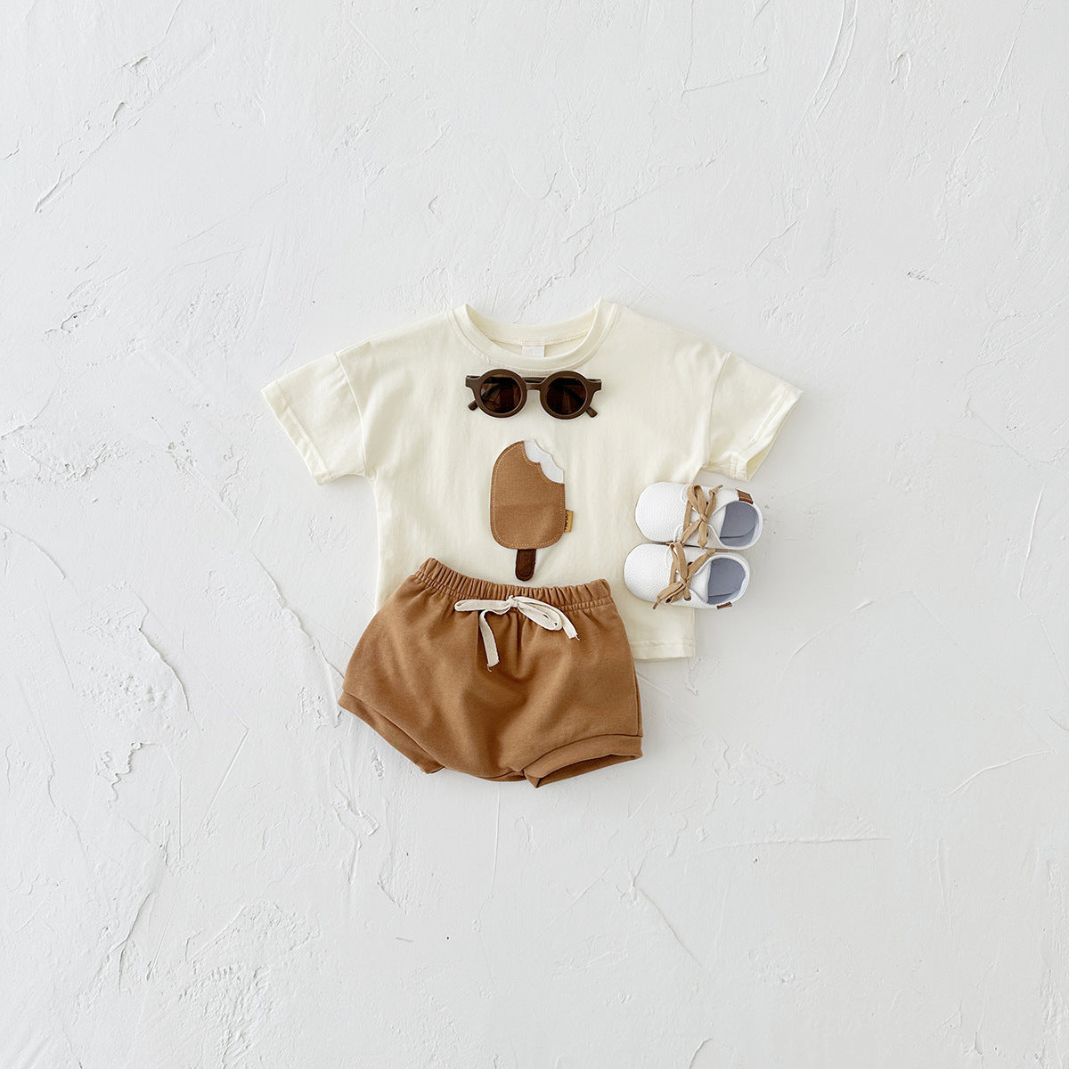 Côte & Crown baby clothes set with cream ice cream print t-shirt, brown shorts, white shoes, and sunglasses