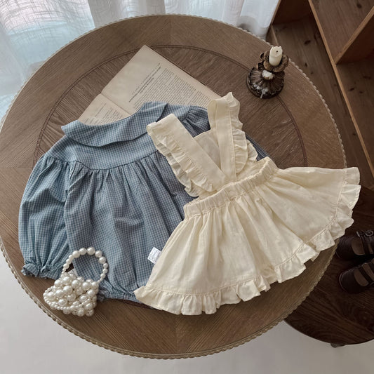 Côte & Crown ruffled baby romper dress in cream with a blue long sleeve romper on a wooden table
